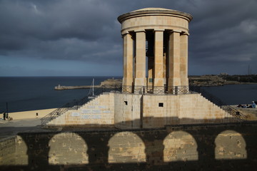 Siege Bell War Memorial beautifully lighted by sunset sunlight, sea, swirling heavy clouds, Valetta, Malta