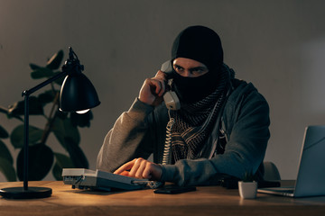Serious criminal in black mask talking on telephone in room