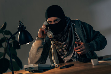 Aggressive terrorist in black mask sitting at table and talking on telephone