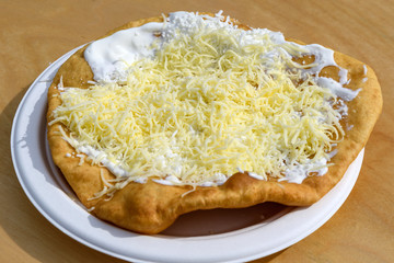 Close up of a langos, typical Hungarian food specialty, with sour cream and cheese on a white dish on a wooden table, deep fried dough in direct sunlight at a food market 