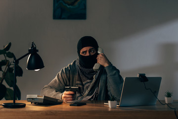 Worried terrorist in mask talking on telephone and holding gun