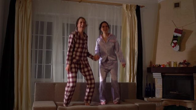 Two happy sisters twins in pajams jumping on the couch in a cozy living room and having fun like in childhood. Relationship sisters. Slow motion.
