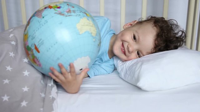 the kid, the boy in the crib, pretends to sleep with the globe in his hands. Child traveler.