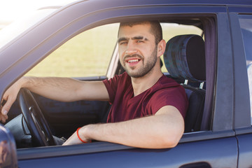 Handsome young dark haired male with stubble dressesd maroon casual t shirt, poses in black car, looks at camera trough window, enjoys his journey, being glad to drive. People, transportation concept.