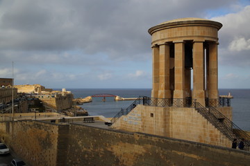 Siege Bell War memorial in valetta, Malta, cars parked on street, historical fortified wall, bridge over sea in background, swirling clouds in sky