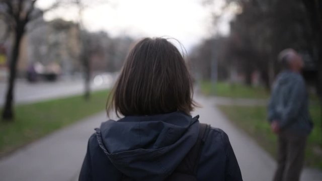 Rear view of casual adult caucasian woman walking down the city street in overcast springtime afternoon, slow motion handheld footage