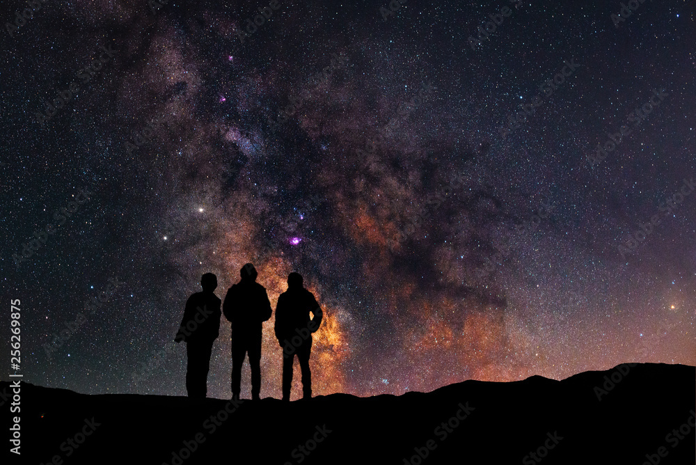 Wall mural three silhouettes stand on a hill and look at the close up milky way galaxy.