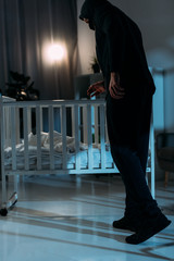 Kidnapper in black clothes standing near crib in dark room
