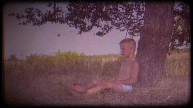 Family video archive. Retro camera 8 mm. Vintage. A little boy sits and dreams under a big tree. Childhood dreams. Place of solitude. Journey back to childhood. Fabulous tree. Memories.  A warm wind