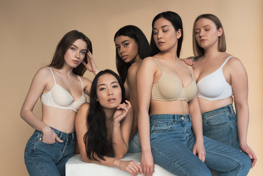 five pretty multicultural girls in blue jeans and bras looking at camera in sunlight, body positivity concept