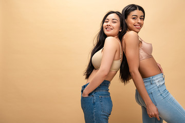 Fototapeta premium two multicultural happy girls in blue jeans and bra standing back to back and looking at camera isolated on beige, body positivity concept