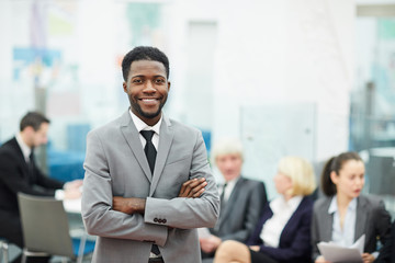 Waist up portrait of successful African-American businessman smiling at camera while posing in office standing with arms crossed, copy space