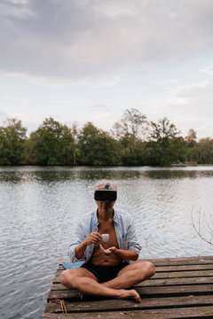 Senior man sitting on jetty at a lake wearing VR glasses and drinking coffee
