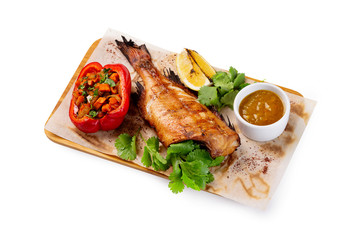 Grilled fish with no head served on paper at wooden board with parsley, pepper, lemon and sauce isolated at white background.