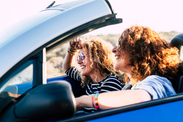 Two curly beautiful women friends drive and travel together on a convertible blue car having fun - outdoor happy leisure activity for cheerful people under the sun of summer - focus on second girl