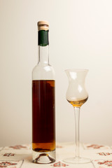 Vertical photo of tall thin bottle of amber colored fortified wine with tulip shaped cordial glass set on embroidered doily