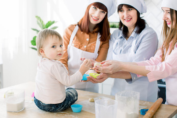 Mothers Day, family, cooking and people concept - little cute baby girl and her mom, aunt and grandmother making and decorating cupcakes at home kitchen