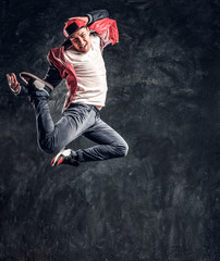 Fototapeta na wymiar Emotional stylish dressed guy performing break dance jumping. Studio photo against a dark textured wall
