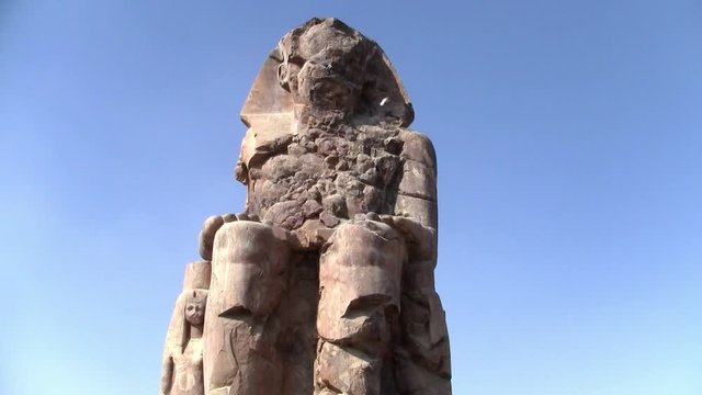 A Memnon Colossus against blue sky. The Colossi of Memnon - Arabic: el-Colossat or es-Salamat - are two massive stone statues of the Pharaoh Amenhotep III located in the Theban Necropolis in Luxor.