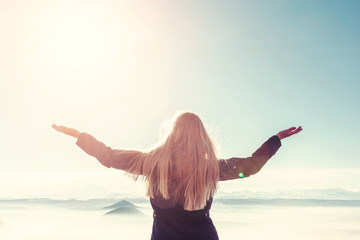 A young blond woman with raised hands enjoys the fresh mountain air and beautiful views of the mountains.