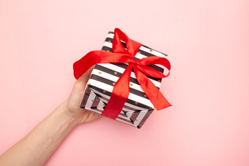 Fototapeta na wymiar woman hands with Gift present box in white and black stripes with a red ribbon bow isolated on pink table top view. Minimal flat lay composition for sales, birthday, mother day or christmas. copyspace
