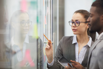 Side view of businesswoman collaborating with colleagues planning startup project placing sticky notes on glass wall, copy space