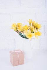 Jar with narcissuses and a cardboard gift box at the white brick background. Spring, mothers day and Easter concept