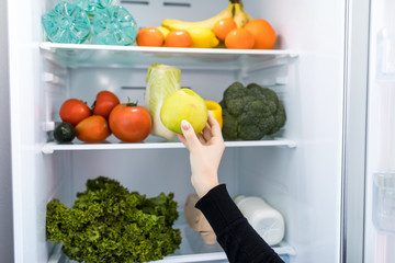 Fresh green vegetables and fruits in fridge. Woman takes the green apple from the open refrigerator. Healthy food.