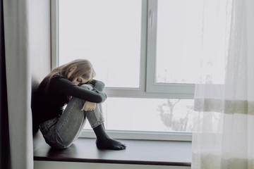 Sad depressed young teenage woman having social problems sitting on windowsill in embryonal position