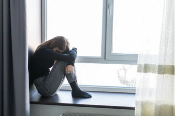 Sad depressed young teenage woman having social problems sitting on windowsill in embryonal position