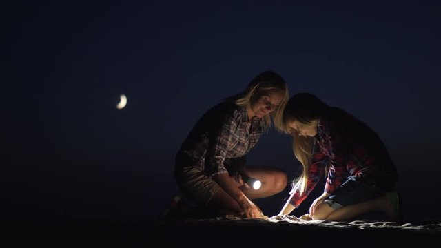 Mom and daughter are playing together in the sand at night. They shine with a flashlight looking for something. Adventure and a happy time together