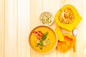 Homemade autumn hot pumpkin cream soup with smocked bacon and seeds. Raw pumpkin, fresh chives, dill, salt in spoon on wooden background, top view, close up.