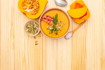 Homemade autumn hot pumpkin cream soup with smocked bacon and seeds. Raw pumpkin, fresh chives, dill, salt in spoon on wooden background, top view.