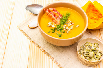 Homemade autumn hot pumpkin cream soup with smocked bacon and seeds. Raw pumpkin, fresh chives, dill, salt in spoon, vintage linen cloth on wooden background, close up.