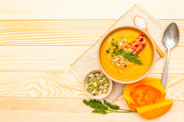 Homemade autumn hot pumpkin cream soup with smocked bacon and seeds. Raw pumpkin, fresh chives, dill, salt in spoon, vintage linen cloth on wooden background, top view, copy space.