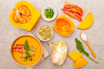 Fototapeta na wymiar Homemade autumn hot pumpkin cream soup with smocked bacon and seeds. Raw pumpkin, bun, fresh chives, dill, salt in spoon, vintage linen cloth on stone background, top view, close up