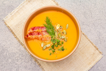 Homemade autumn hot pumpkin cream soup with smocked bacon and seeds. Raw pumpkin, fresh chives, dill, vintage linen cloth on stone background, top view, close up.