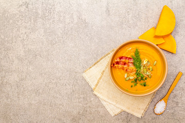 Homemade autumn hot pumpkin cream soup with smocked bacon and seeds. Raw pumpkin, fresh chives, dill, salt in spoon, vintage linen cloth on stone background, top view, copy space.
