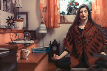 Young woman meditating, exercising yoga in a lotus position at her home, sitting next to a window