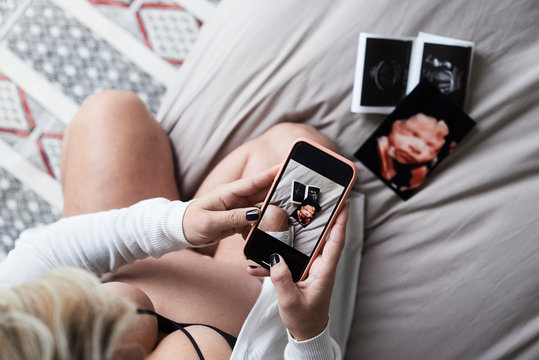 Pregnant woman at home sitting on bed taking pictures of ultrasound images