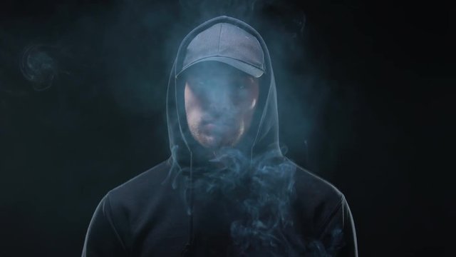 Male bully in hoodie smoking cigarette against dark background, night criminal