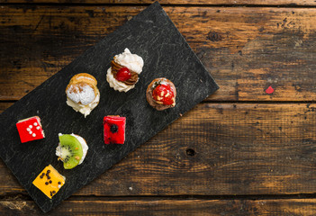 Selection of different dessert pastry sweet mini cakes and tartlets with cream and fruit on slate and wooden background