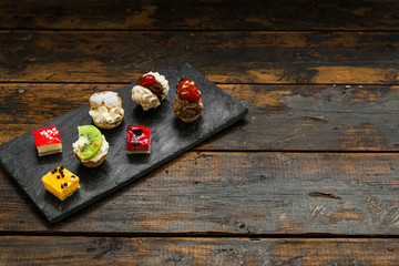 Selection of different dessert pastry sweet mini cakes and tartlets with cream and fruit on slate and wooden background