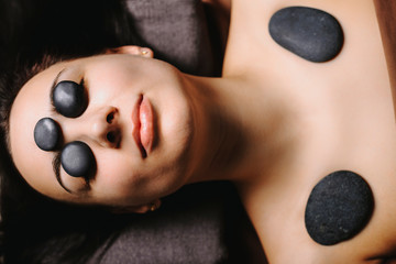 Upper view of a beautiful adult female with dark hair resting on a spa bed with closed eyes and with hot volcanic stones on her face and body ina spa salon while traveling.