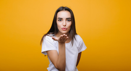 Obraz premium Portrait of a cute european female looking at camera and doing a air kiss while dressed in white shirt in front of a yellow studio background.