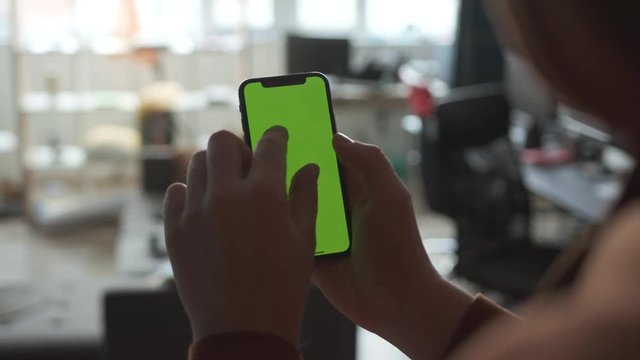 Man's hand uses mobile smartphone with green screen in vertical position indoors in the living room. Tapping the screen, scrolling pages, updates. Communication, messaging. Close up view