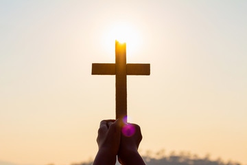 silhouette human hands praying to the GOD while holding a crucifix symbol with bright sunbeam on the sky