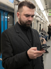 Handsome bearded man dressed in wool coat stands in metro carriage and uses a smartphone