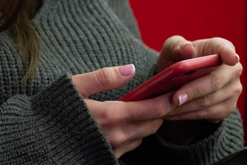 Red smartphone in girlish hands. The girl in a knitted sweater holds a mobile phone. Close-up.