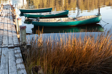 Three wooden boats tied to an old wooden jetty, bob in the water in the golden light of the evening.